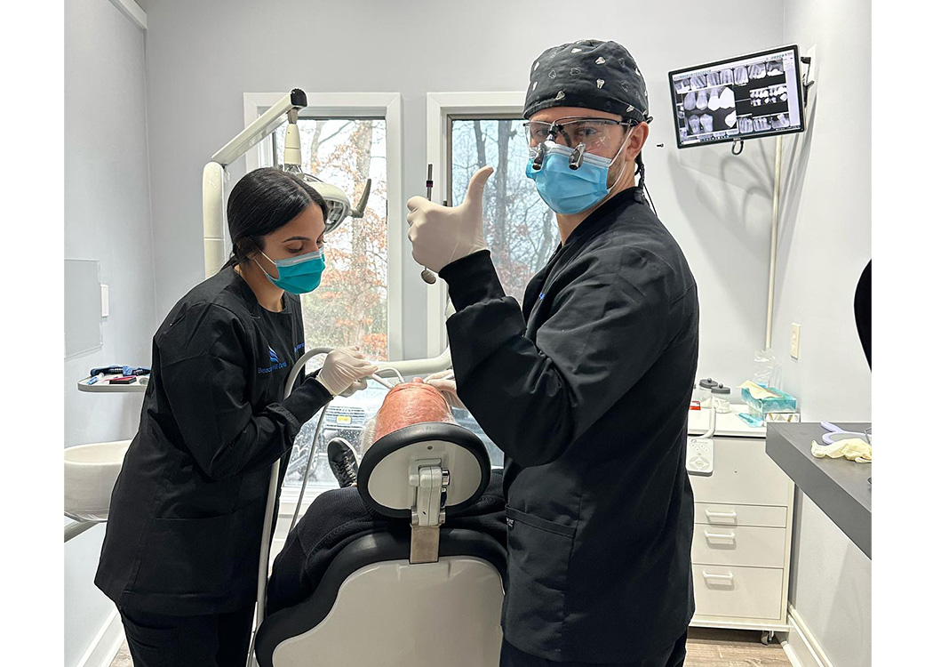 The image shows two dental professionals in a dental practice, with one person sitting in the dentist s chair and another standing behind them, both wearing personal protective equipment.