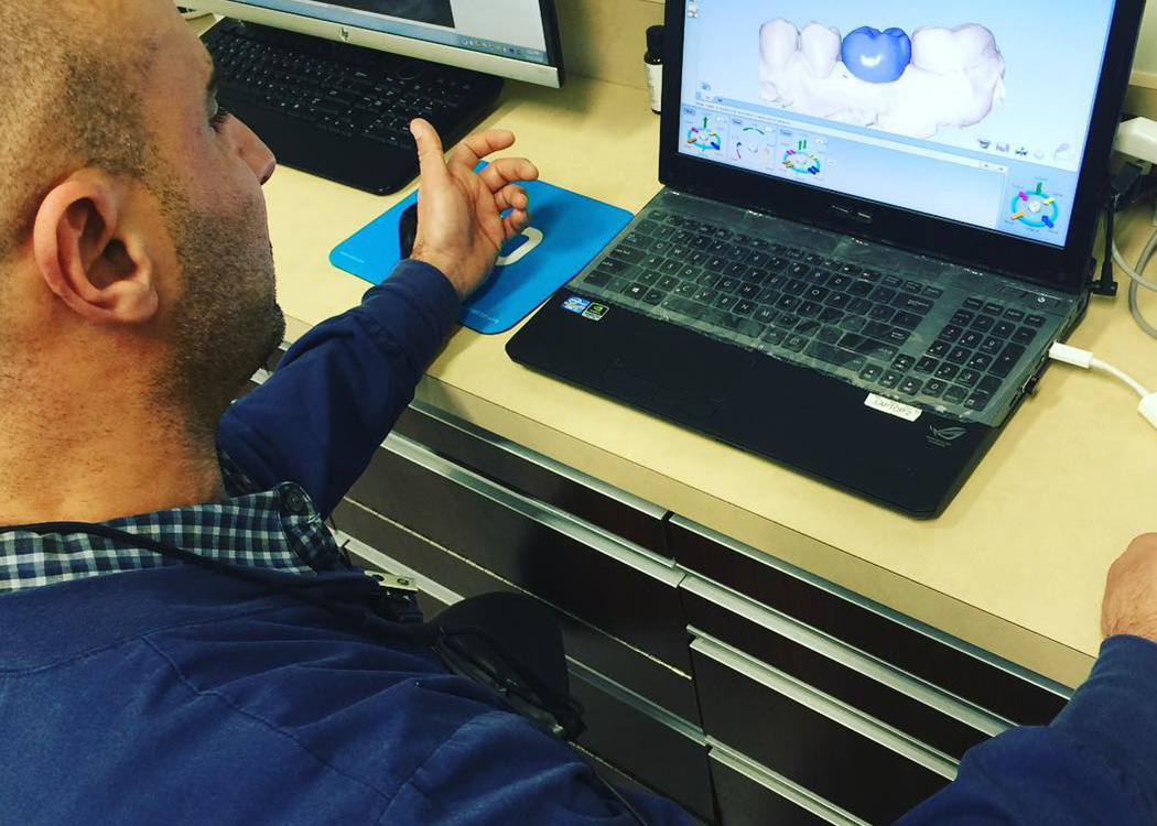 The image shows a man using a laptop with a 3D model displayed on the screen, which appears to be related to dental or medical technology. He is seated at a table with various equipment and tools around him.