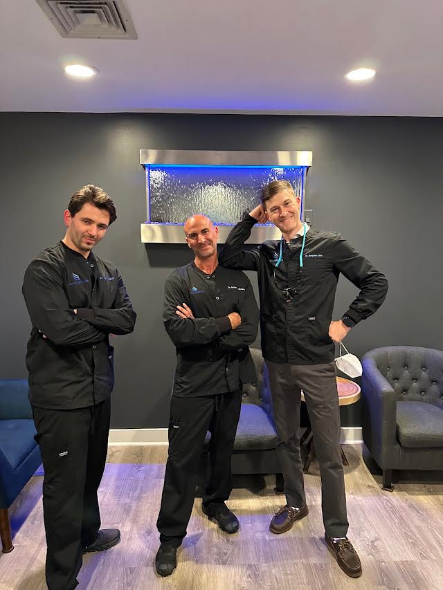 Three men in medical scrubs posing for a photo with their arms crossed, standing in front of a couch and a wall-mounted sign.