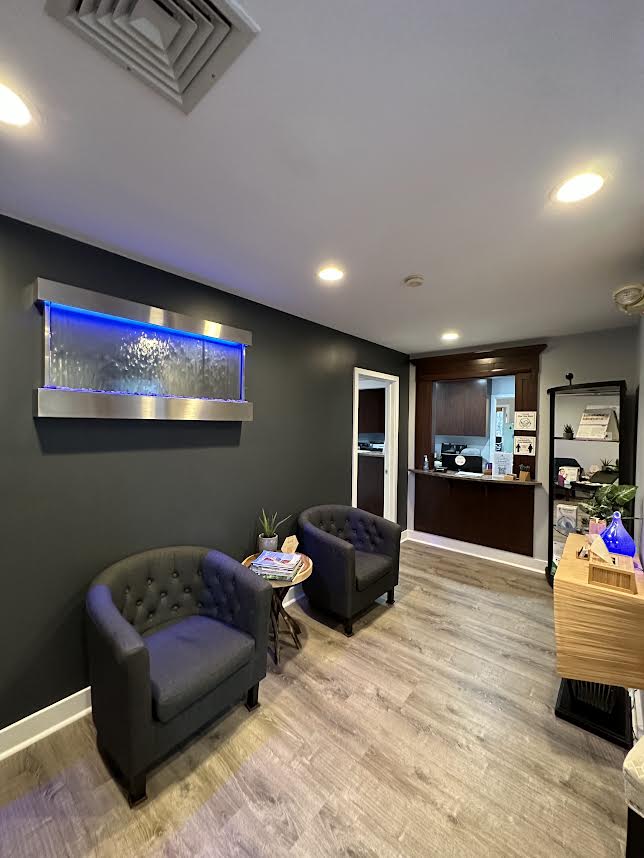 The image shows an interior space that appears to be a waiting area or lobby, with dark walls and flooring, a blue wall-mounted light fixture, a black leather sofa, a wooden desk, and a receptionist behind it. There is a large window with a white shade partially drawn.