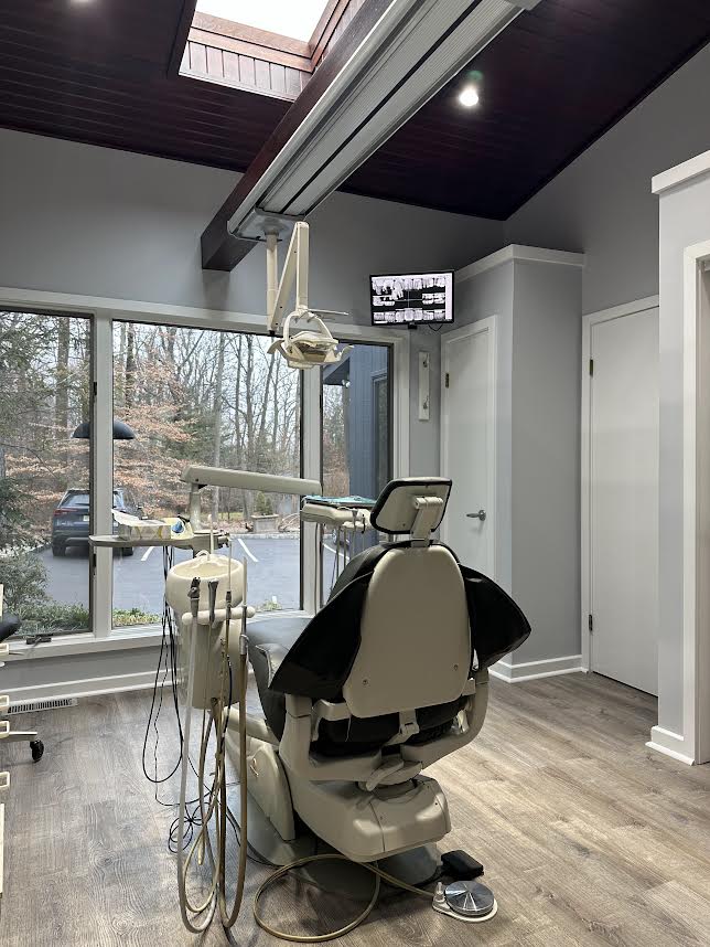 The image shows a dental office interior with a large dental chair, a computer monitor displaying what appears to be an X-ray or dental imaging, and equipment typically found in a dental practice.