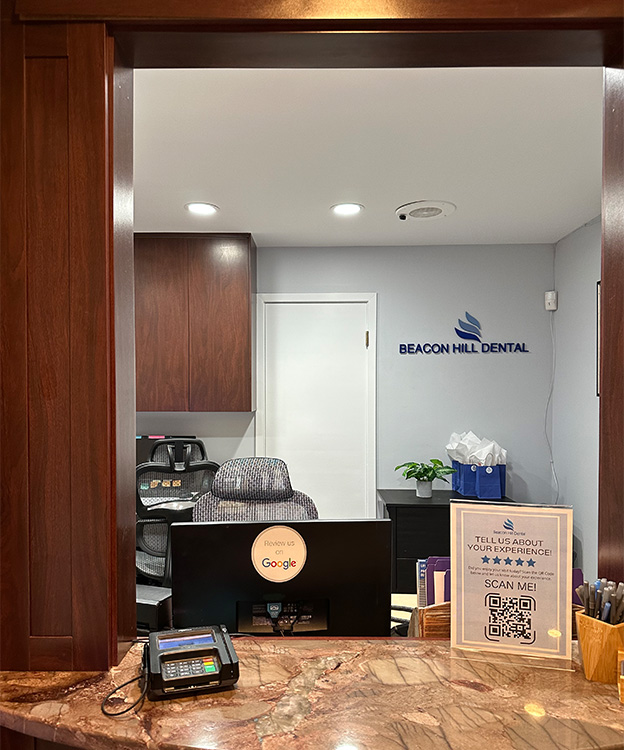 The image shows an interior view of a dental office reception area, featuring a modern design with a large window, a desk with a computer monitor and keyboard, a blue sign with the name  Beacon Hill Dental  on the wall, a receptionist area, and a waiting room.