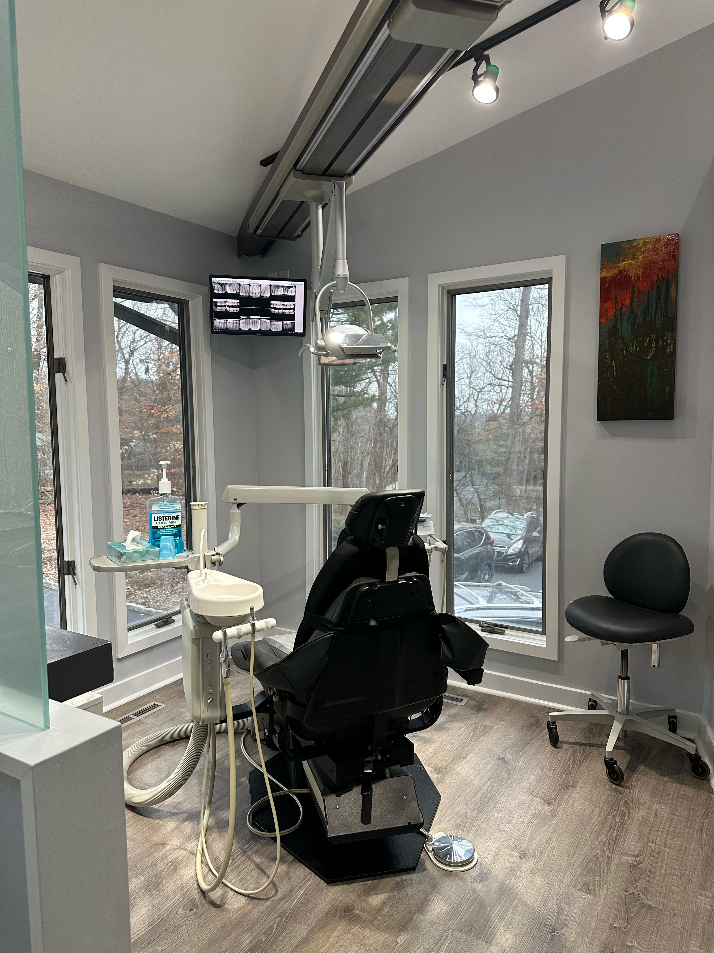 The image shows a dental office interior, featuring a dental chair with a black leather seat and headrest, a modern dental equipment setup including a dental light and an adjustable dental chair, a large window allowing natural light to enter the room, and various pieces of dental equipment such as a dental mirror, dental instruments, and a computer screen displaying patient information. The office is well-lit with recessed ceiling lights and has a clean, professional appearance with white walls and flooring.