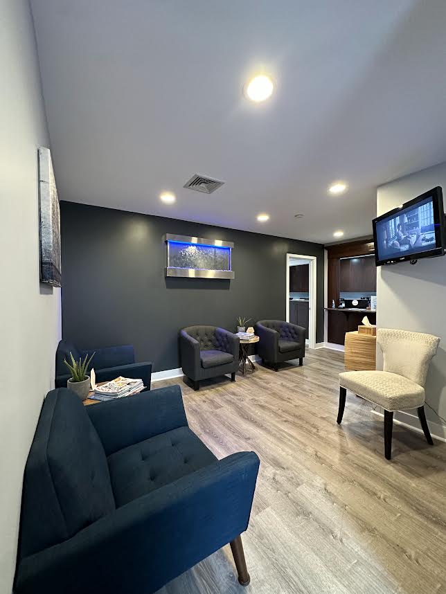 The image shows a well-lit, modern waiting room with a comfortable seating area, a television mounted on the wall, and a reception desk.