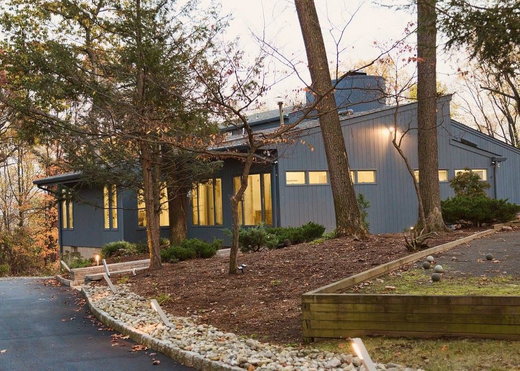 An image depicting a modern, two-story house with a gray exterior and large windows, situated in a residential area with trees and a driveway leading up to it. The sky is overcast.