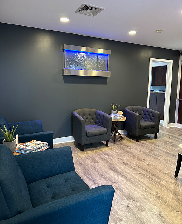 The image shows an interior space of a waiting area with a modern design, featuring blue walls, a dark ceiling, a gray wall-mounted panel displaying a digital art piece, a blue sofa and chair, and a large window with a black frame.