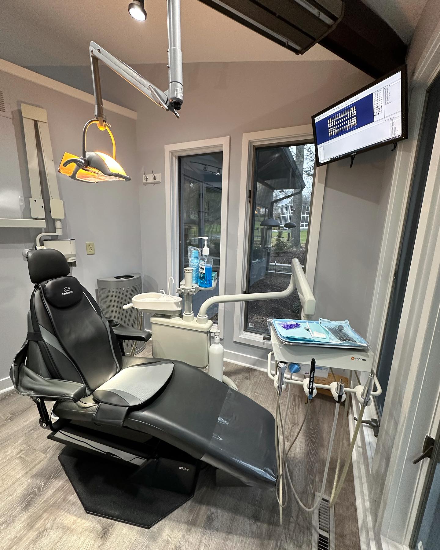 The image shows an interior view of a dental office with a chair and equipment, including monitors displaying patient information.