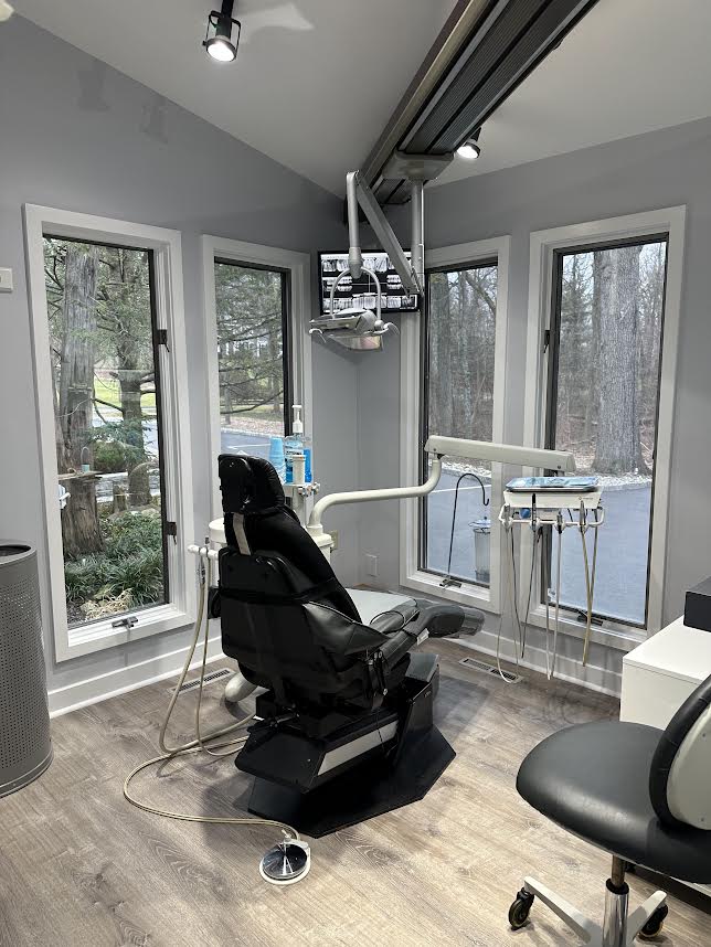 The image shows an interior view of a dental office with a chair, a desk area with a computer monitor and keyboard, dental equipment, and a window with natural light.