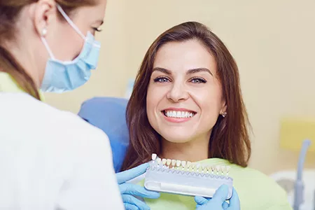 A dental professional smiling at a patient in a dental chair, with the dentist holding a dental device and preparing to work on the patient s teeth.