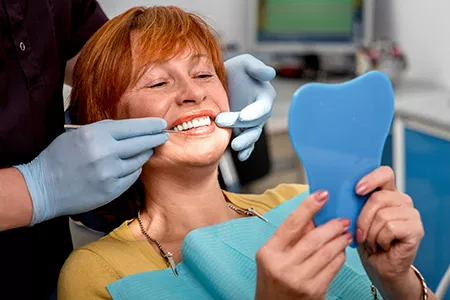 A woman is seated in a dental chair, smiling broadly, with her teeth being attended to by a dentist who is holding up a mirror for inspection.