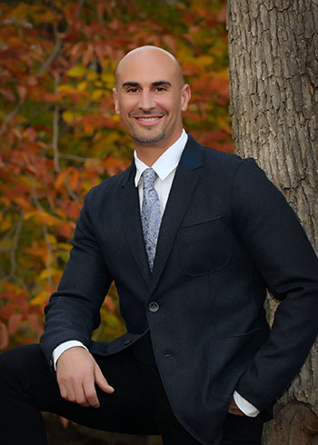 A man in a suit and tie is standing in front of a tree with autumn foliage, smiling at the camera.