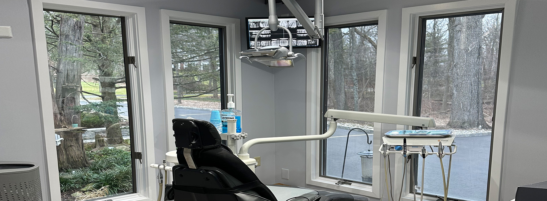 The image shows an interior view of a dental office, featuring a reception area with a window and a desk, a waiting area with chairs and a TV screen on the wall, and a dental treatment room with a chair, a light fixture, and a counter.