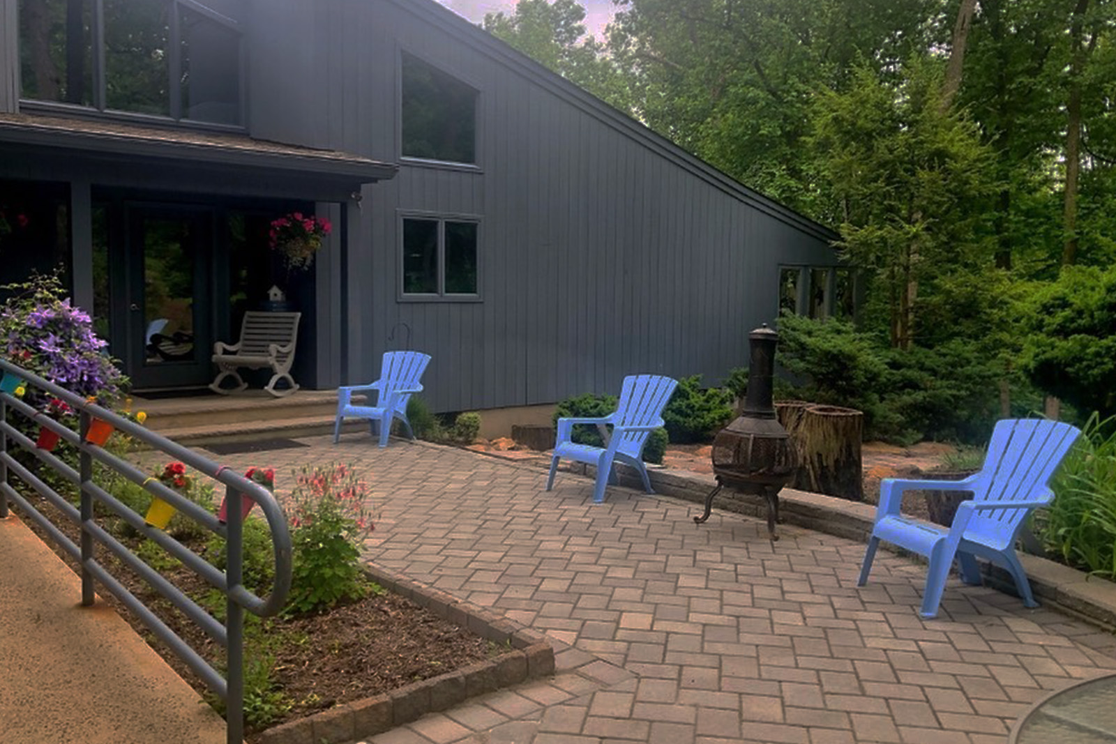 The image shows a backyard patio area with outdoor furniture, including chairs and a dining table, set on a brick pavement. There is a wooden deck with a railing leading to the patio, and a blue bench is placed near a fire pit. A house with a dark exterior and windows is visible in the background. The sky is overcast, and there are trees surrounding the property.