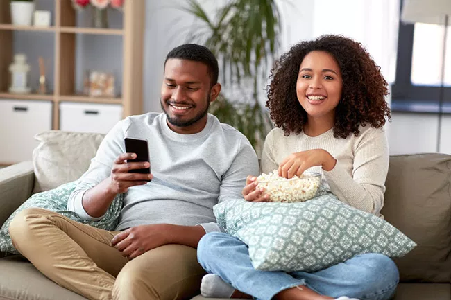 Two individuals, a man and a woman, seated on a couch, sharing a moment of relaxation with snacks while engrossed in content on a smartphone.