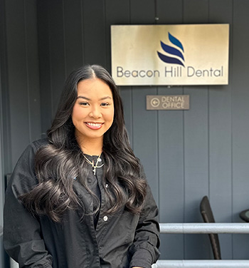 The image shows a person standing in front of a sign that reads  Beacon Hill Dental Office.  The individual is smiling and posing for the photo.