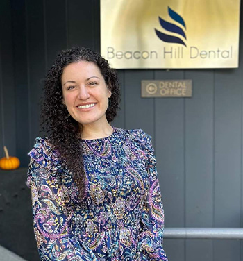 A woman stands in front of a sign for  Beacon Hill Dental Office,  smiling at the camera.