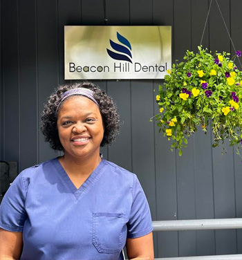 The image shows a woman standing in front of a sign that reads  Beacon Hill Dental.  She is wearing scrubs and has a smile on her face.