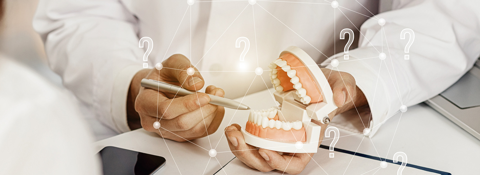 The image shows a person s hand holding a dental model with teeth and gums, while another person is seen in the background working at a computer.