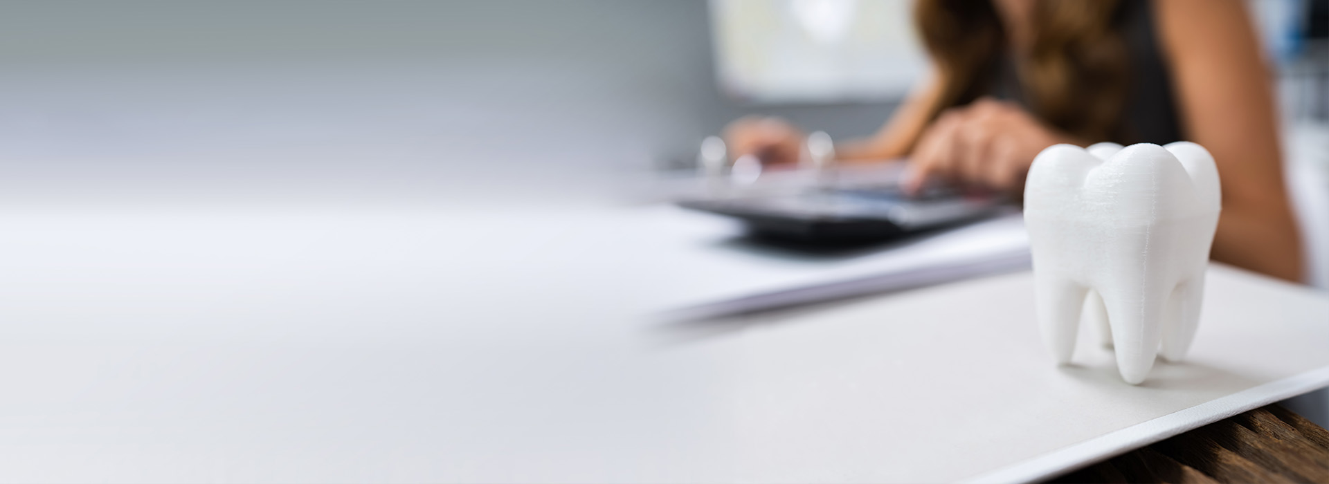 The image features a person working at a desk with a computer monitor, keyboard, and mouse visible. In the foreground, there is a white object resembling a tooth or a small sculpture placed on a surface to the right of the monitor. The background is blurred but shows an indoor setting with natural light coming in from the left side of the frame.