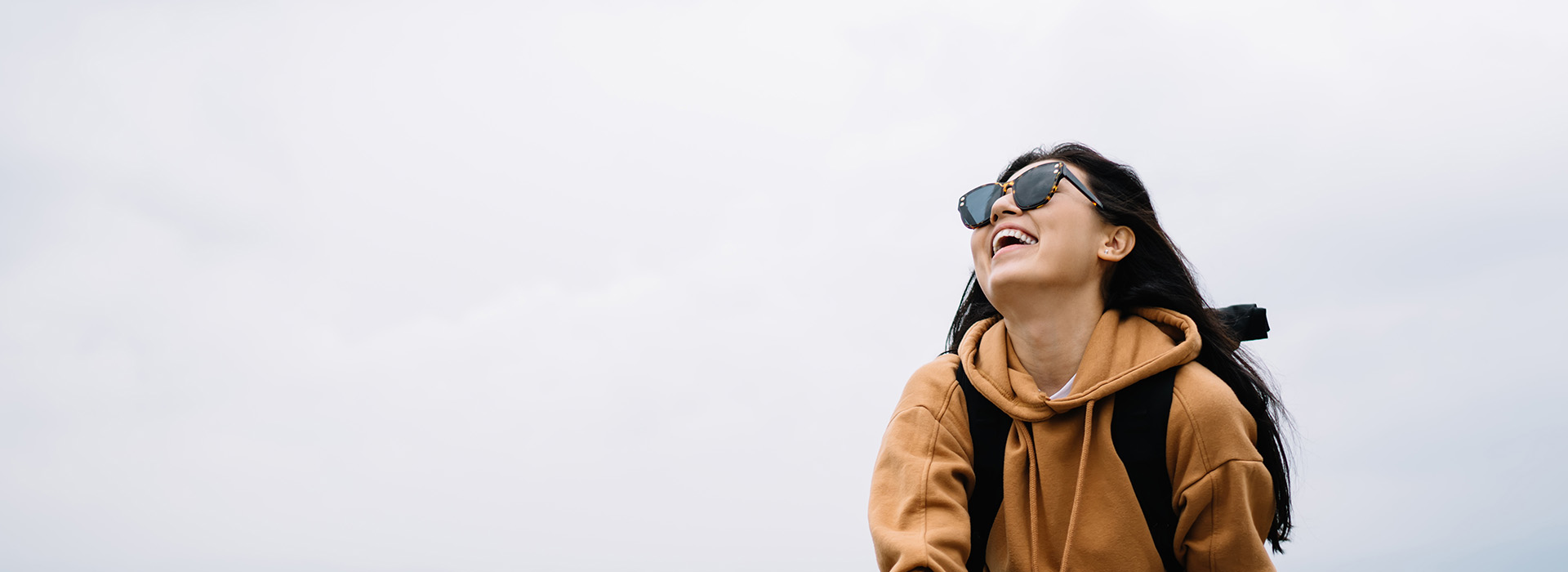 A person in a beige hoodie and sunglasses, smiling and looking upwards, against a cloudy sky background.