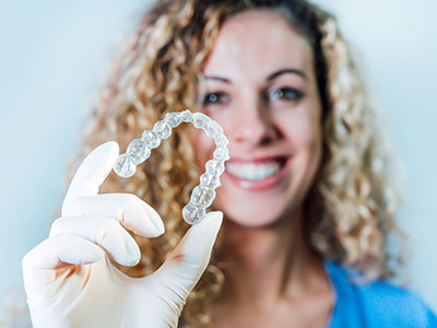 The image features a woman holding a clear dental retainer, smiling and wearing a blue shirt.