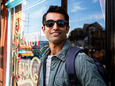 The image shows a man standing in front of a storefront, wearing sunglasses and a backpack, with a friendly expression.