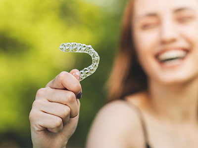 A smiling woman holding a clear plastic aligner, showcasing its use for dental orthodontics.