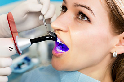 The image shows a woman receiving dental treatment, with a dental hygienist using a device to clean her teeth while wearing protective gloves and a mask.