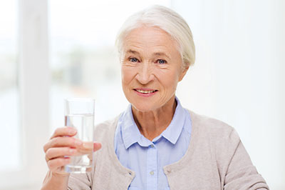 The image shows an older woman holding a glass of water, smiling and looking directly at the camera.