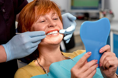 A woman in a dental office, holding a blue dental model while smiling and looking at the camera. She is wearing an orange headband and has a toothbrush around her neck.