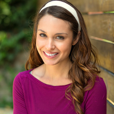 The image is a portrait of a smiling woman with long hair, wearing a purple top and a headband, posing against a wooden fence.