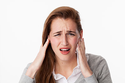 The image shows a woman with her hand on her head, possibly in pain or discomfort. She appears to be indoors and is wearing a grey cardigan.