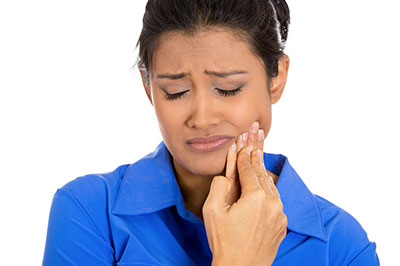 The image shows a woman with her eyes closed and her hand to her face, possibly in pain or discomfort. She appears to be looking upwards and is wearing a blue top.