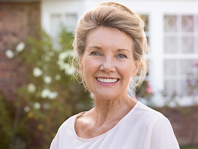 Woman in a cream top with a smile, standing in front of a house with a brick wall and greenery.