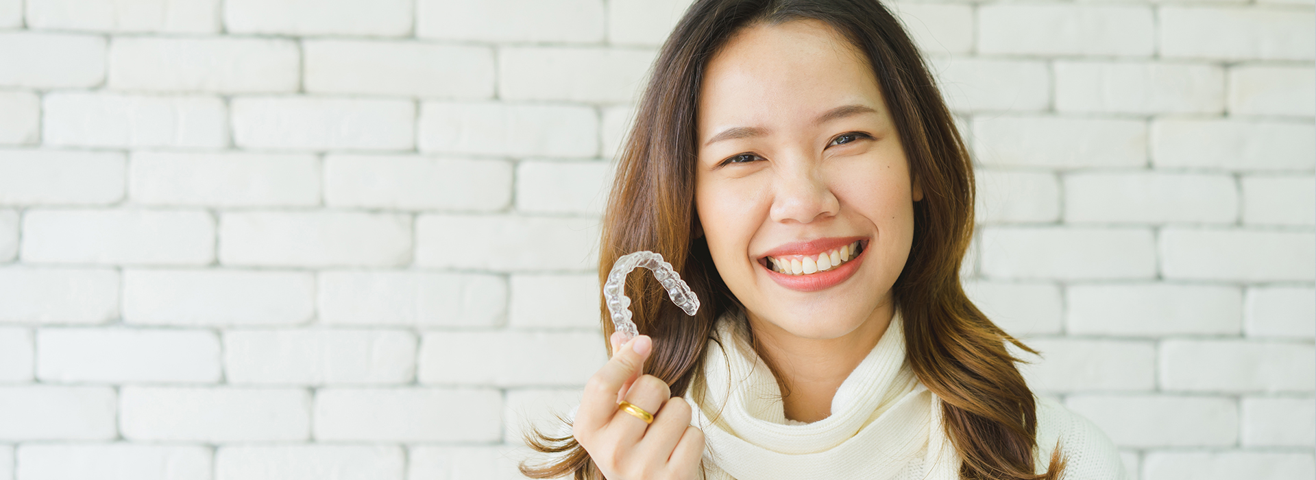 The image shows a smiling woman with dark hair, wearing a white top and holding a ring in her hand. She is standing against a brick wall background.