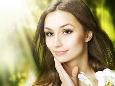 A woman with long, dark hair and fair skin is shown in a close-up. She has a neutral expression and appears to be looking directly at the camera. Her makeup is natural, enhancing her facial features. The background is out of focus but suggests a serene, natural setting with hints of greenery and light filtering through.