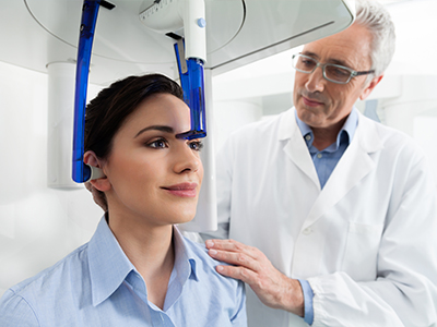 The image depicts a person seated in front of a large, blue 3D scanner machine. A medical professional is standing behind the individual, wearing a white lab coat and glasses, observing the process with interest.