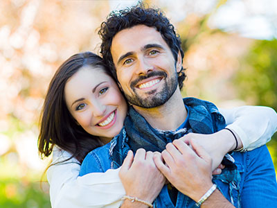 The image shows a man and a woman smiling at the camera, with the man appearing to be in his mid-adulthood and the woman slightly younger. They are standing close together, suggesting a friendly or intimate relationship between them.