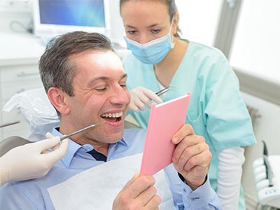 The image features a man in a dental chair, holding a pink card with both hands, smiling at the camera while looking down at the card. He is seated in front of a female dental professional who is also smiling and appears to be engaged in conversation or examination. They are both in a well-lit dental office setting, with medical equipment visible in the background.