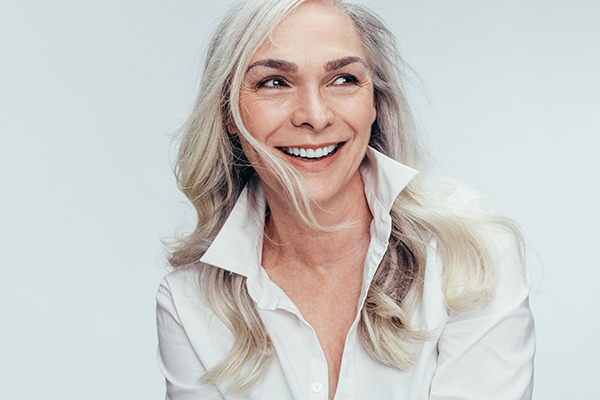 The image is a photograph of a woman with short, light-colored hair smiling at the camera. She has a fair complexion and is wearing what appears to be a white top.