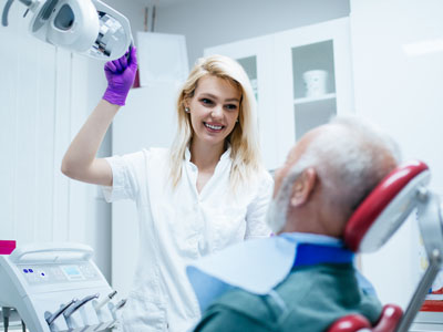 The image shows a dental professional in a white lab coat assisting an elderly patient with a dentist s chair and equipment, with the professional holding up a dental mirror to inspect the patient s mouth.