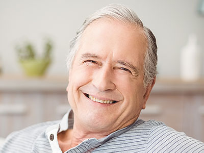 The image shows an older man with a smile, wearing a dark shirt and sitting in a comfortable chair.